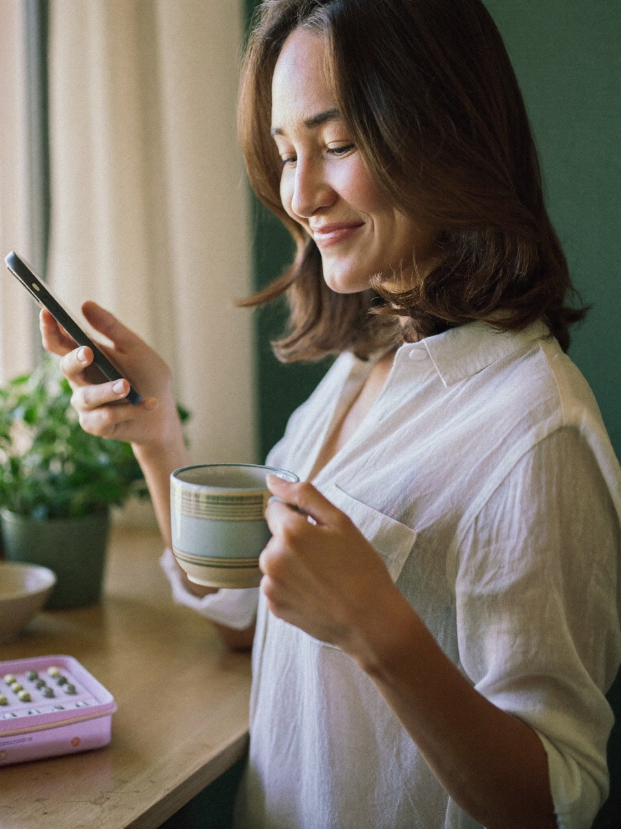 A woman at home in the morning, smiling at her phone with a cup of tea, a lilac pill organiser on the table